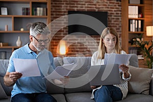 Sad mature caucasian couple work with documents and laptop, pay bills and taxes at evening in living room
