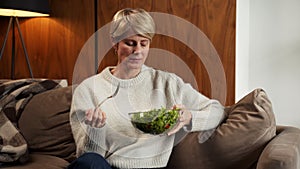 Sad middle-aged woman eats a tasteless green vegetable salad sitting on the couch