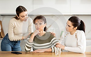 Sad middle-aged woman sitting at table while two others trying to calm her