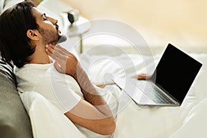 Sad middle eastern guy woke up, sits on bed, check tonsils with laptop with blank screen in bedroom