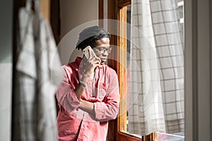 Sad upset African man having difficult conversation on phone, standing by window at home photo