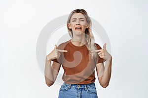 Sad and miserable girl pointing fingers at herself while crying, sobbing, standing against white background