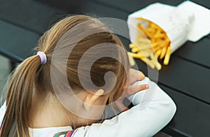Sad, moody girl doesn`t want to eat French fries. Tired cute little girl tired of sitting at a dark wooden table in a cafe. Angry