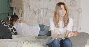 Sad nervous woman worried by problems in the relationship sitting in bed, her boyfriend is lying near turning away.