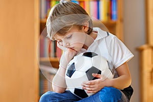 Sad and not happy little kid with football about lost football or soccer game. child after watching match on tv