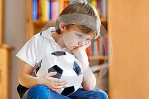 Sad and not happy little kid with football about lost football or soccer game. child after watching match on tv