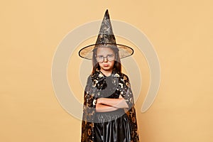 Sad offended female kid girl wearing witch black hat costume isolated over beige background expressing sorrow and sadness keeps