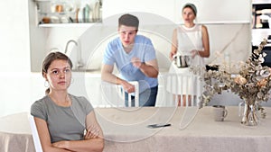 Sad offended young woman sitting at kitchen table during argument with dissatisfied husband and mother-in-law standing