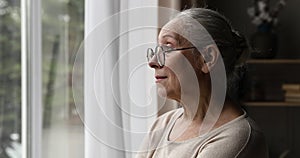 Sad older woman sighing stands near window and looking outside