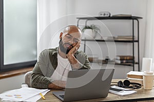Sad overworked latin man sitting at workdesk in front of laptop at home office and looking at screen, free space