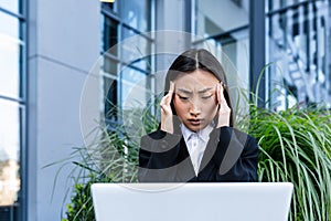 Sad and pensive business woman working on laptop sitting on a bench near the office, Asian tired and depressed looking for work