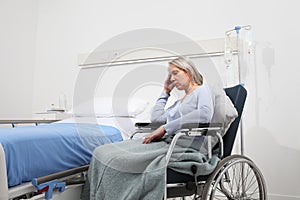 Sad and pensive elderly woman abandoned and forgotten isolated on wheelchair near bed in hospital room, concept of loneliness and