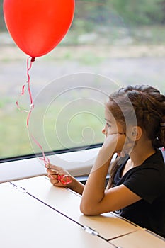 Sad pensive little girl with the red balloon in her hand looking out the train window