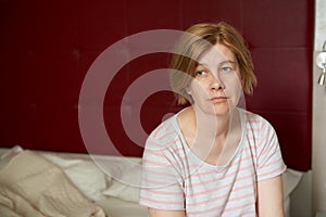 A sad, pensive middle-aged mature woman sits on a bed in a bedroom.