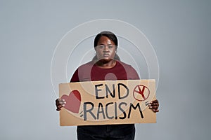 Sad plus size young african american woman in casual clothes looking at camera, holding End Racism banner in front of