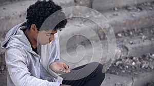 Sad poor teenager looking at coins in hand sitting outdoors, urban unemployment