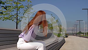 Sad pretty red-haired girl wearing casual clothes sitting on bench in city park on bright sunny day.