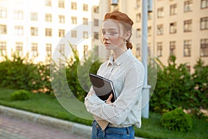 A sad red-haired girl with freckles on her face holds a folder in her hands.