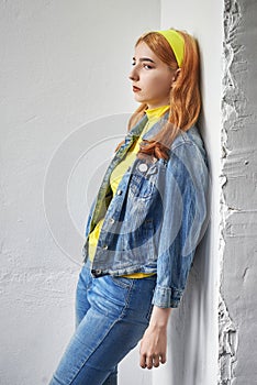 Sad red-haired young girl in yellow and blue clothes stands against the wall