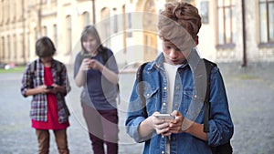 Sad redhead Caucasian schoolboy using smartphone, sighing. and looking back at classmates teasing him. Blurred offenders