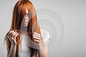 Sad redhead girl holding her damaged hair looking at camera.