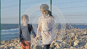 Sad refugee children stand on the beach near border. Restricted area net fence protected region. Sorrowful boy and girl stand alon