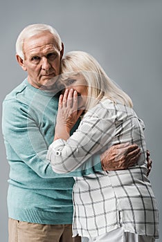 sad retired man hugging frustrated senior wife covering face isolated on grey.