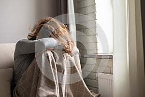 Sad scared woman in panic attack covering her face with hands and crying. Lonely girl wrapped in a plaid sitting near window