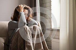 Sad scared woman in panic attack grabbing her head by hand. Lonely girl wrapped in a plaid sitting near window of apartments.