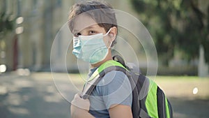 Sad schoolboy with backpack in face mask turning to camera. Portrait of upset Caucasian boy standing on sunny schoolyard