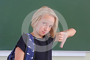 Sad schoolgirl with a backpack is showing a thumbs down in a classroom near green chalkboard. The child does not like the school.