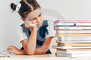 Sad schoolgirl looking on a pile of books on the desk against light grey background. Exhausted little girl learner has tired