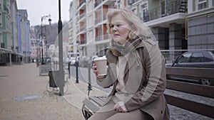 Sad senior Caucasian woman sitting on bench outdoors and drinking coffee. Lonely elegant retiree resting on city street