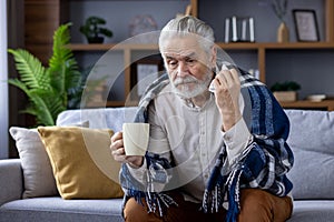 Sad senior gray-haired man sitting on sofa at home and suffering from illness. Covered with a blanket, holding a cup of