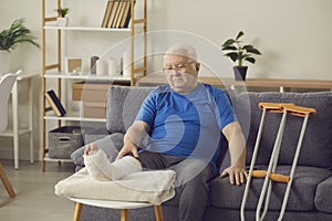 Sad senior man with broken leg sitting on sofa at home and thinking about his injury