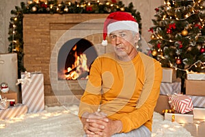 Sad senior man sitting in living room and looking at camera, celebrating christmas eve alone, has upset facial expression, wearing
