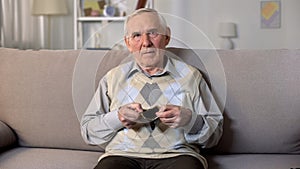 Sad senior man sitting on sofa showing empty wallet on camera, credit poverty