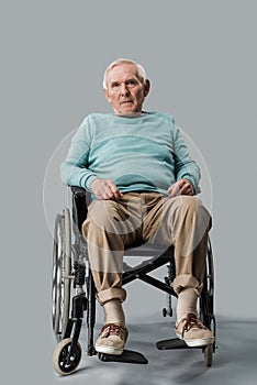 sad senior man sitting in wheelchair and looking at camera on grey.