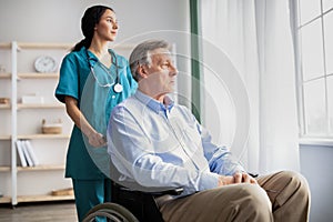 Sad senior man in wheelchair feeling depressed, looking out window, young nurse helping him at home