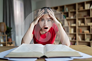 Sad shocked teen chinese lady student with open mouth study at table with book in room or library interior