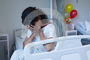 Sad sick mixed race girl sitting on hospital bed crying, with head in hands