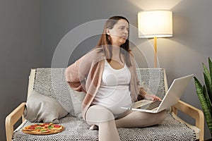 Sad sick tired pregnant businesswoman professional sitting on sofa having backpain while working online on laptop grimacing from