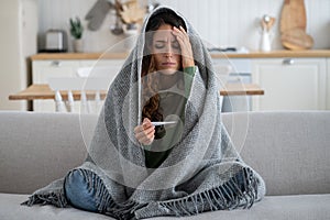 Sad sick woman with warm blanket sits on sofa and looks at thermometer waiting for arrival of doctor