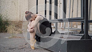 Sad slim Caucasian ballerina in black dress sitting outdoors hugging knees. Portrait of exhausted upset young woman