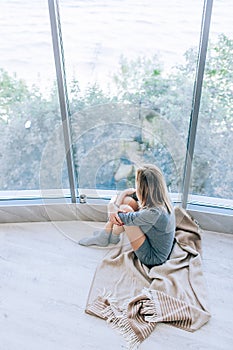 Sad slim woman sitting on a warm floor in a grey dress near the large window in light and hugging her knees. Autumn mood, warmth