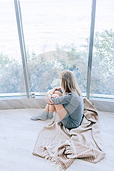 Sad slim woman sitting on a warm floor in a grey dress near the large window in light and hugging her knees. Autumn mood, warmth