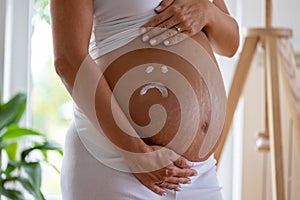 Sad smiley drawn with cream on pregnant belly with stretch marks