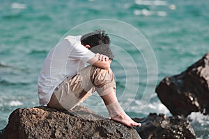 Sad stressed young Asian man hug his knee sitting on the rock of sea shore