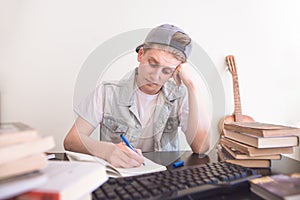 Sad student sitting at the table near the books and computer and writing a pen in a notebook