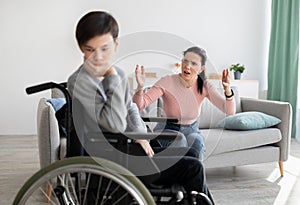 Sad teen boy in wheelchair having conflict with his angry mother, listening to her screaming at home